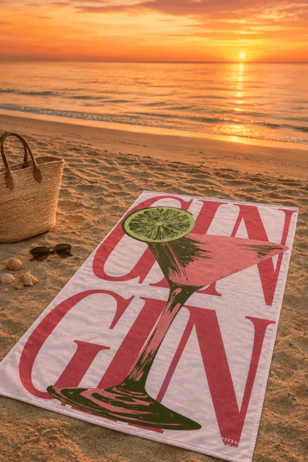 Demi Bravo GIN Coctail plaj havlusu, gün batımında kumsalda hasır çanta, güneş gözlüğü ve deniz kabuklarıyla birlikte serili.