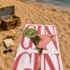 GIN Coctail plaj havlusu kumsalda piknik sepeti, içecekler, kitap ve güneş gözlüğüyle serili; denizde bir yelkenli görülüyor.