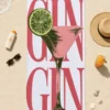 Kokteyl kadehi ve lime desenli Demi Bravo GIN Coctail plaj havlusu, deniz kıyısında şapka, güneş gözlüğü ve içecekle çevrili.