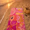 Pink Summer Plaj Havlusu, pembe ve turuncu tonlardaki narenciye, balık, papaya, leopar, çiçek ve denizyıldızı desenleriyle gün batımında kumsala serili.