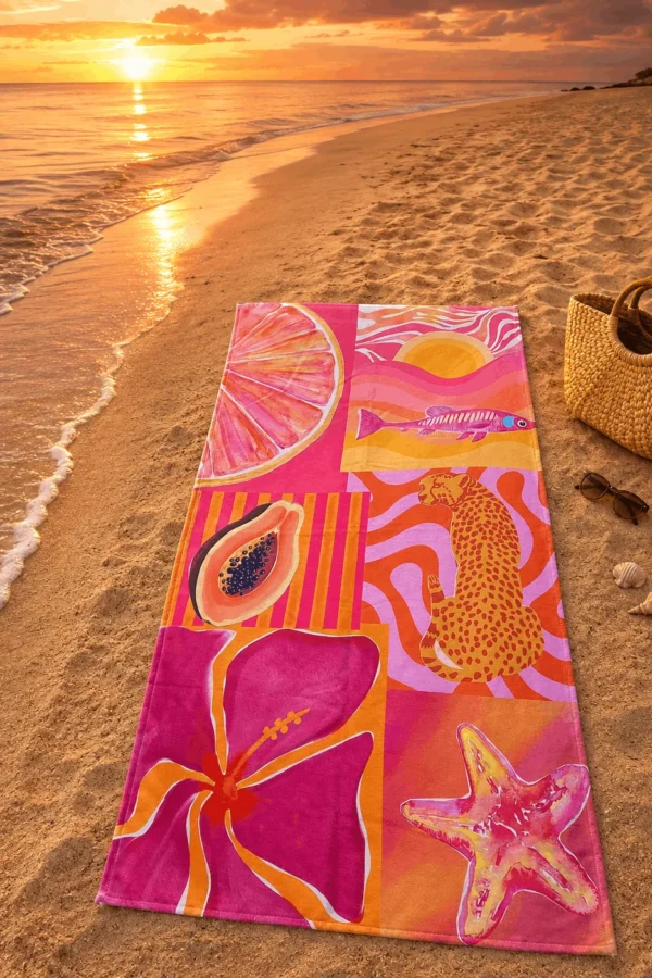 Pink Summer Plaj Havlusu, pembe ve turuncu tonlardaki narenciye, balık, papaya, leopar, çiçek ve denizyıldızı desenleriyle gün batımında kumsala serili.