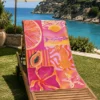 Pink Summer Plaj Havlusu, pembe ve turuncu hayvan, meyve ve deniz temalı desenleriyle deniz manzaralı bahçedeki ahşap şezlong üzerine serilmiş.
