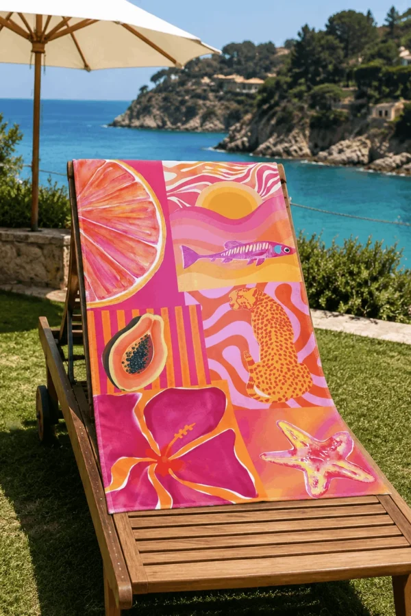 Pink Summer Plaj Havlusu, pembe ve turuncu hayvan, meyve ve deniz temalı desenleriyle deniz manzaralı bahçedeki ahşap şezlong üzerine serilmiş.