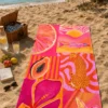 Pink Summer Plaj Havlusu, pembe ve turuncu leopar, papaya, balık, çiçek ve denizyıldızı desenleriyle kumsalda piknik sepetinin yanında serili.
