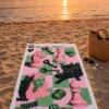 Pop Art Chess Plaj Havlusu, pembe ve yeşil dama zemin üzerindeki satranç taşları, anahtarlar, zarlar ve siyah kedilerden oluşan deseniyle gün batımında kumsala serili.