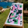 Pop Art Chess Plaj Havlusu, pembe satranç taşları, siyah kediler ve yeşil zar desenleriyle deniz manzaralı bahçedeki ahşap şezlong üzerine serilmiş.