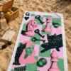 Pop Art Chess Plaj Havlusu, satranç taşları, siyah kediler, anahtarlar ve zar desenleriyle deniz kıyısındaki kum üzerine serili; yanında piknik sepeti ve içecekler bulunuyor.