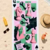 Pop Art Chess Plaj Havlusu, pembe ve yeşil kareli zemindeki satranç taşları, siyah kediler, anahtarlar ve zar desenlerini gösteren üstten görünümle kum üzerinde serili.