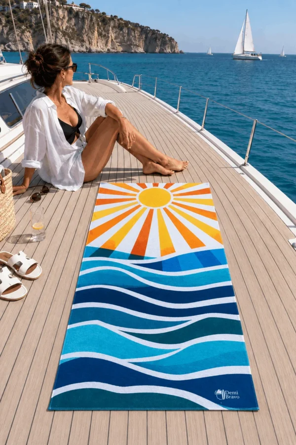 Sun Plaj Havlusu, sarı ve turuncu güneş ışınları ile mavi tonlardaki dalga desenleriyle gün batımında deniz kıyısındaki kuma serili.